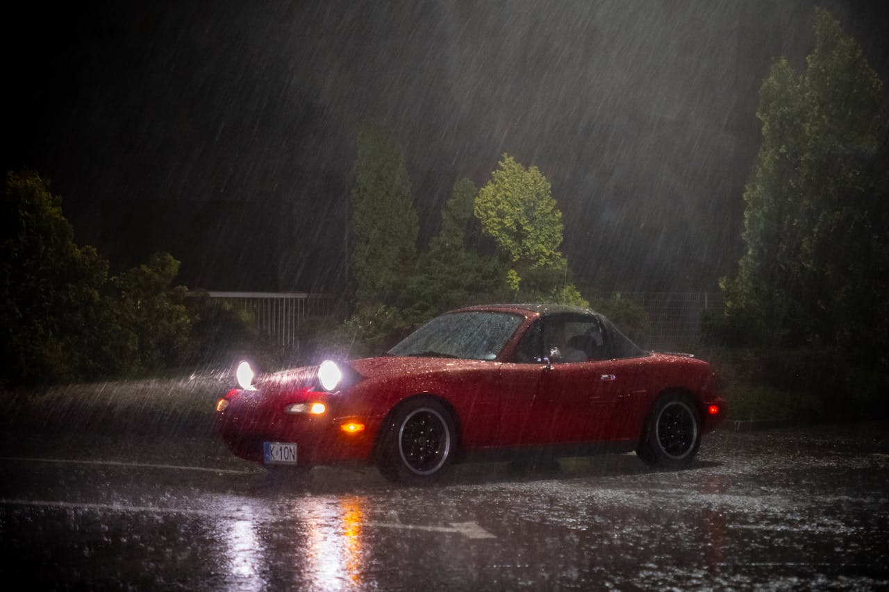 Red Mazda Miata parked in heavy rain at night, showcasing vehicle lights and wet roads.